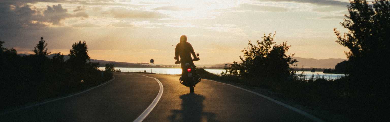 Motociclista viajando numa estrada asfaltada, com arbustos nas laterais e um lindo pôr do sol.